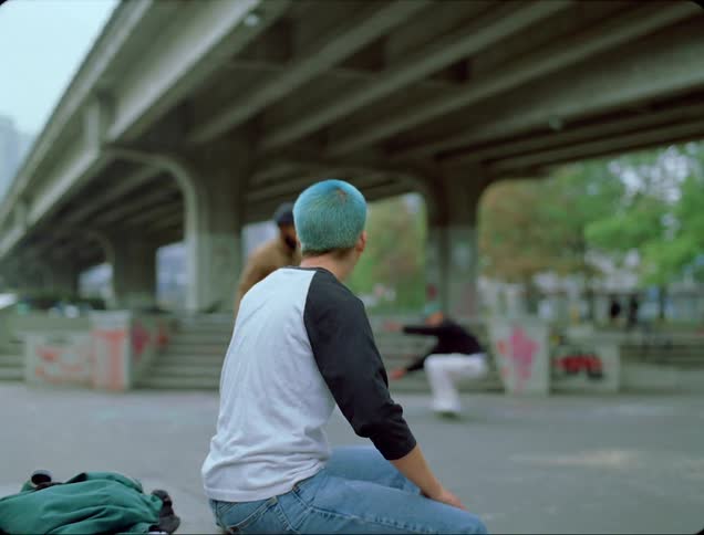 One of several people skateboarding under overpass near city downtown passes by sitting woman and high fives her