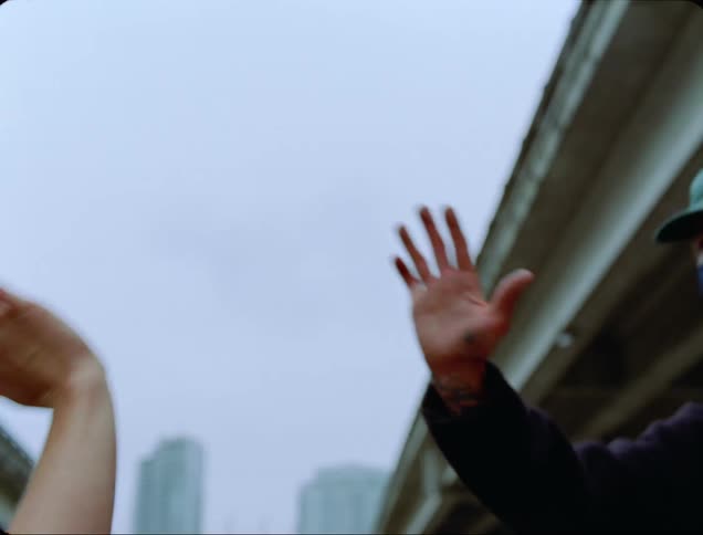 A person with a hat and a mask underneath an overpass with buildings in the background