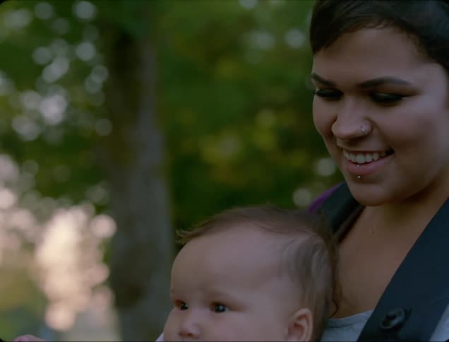 A woman carries her baby in a front carrier and smiles as they stand by their vehicle in a park