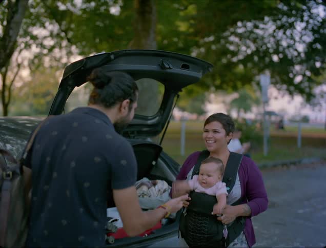 A man in a parking lot at a public park walks up and greets a woman and her baby
