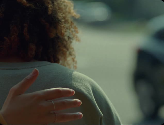 Cars drive past as one woman has her hand behind another womans back as she sits