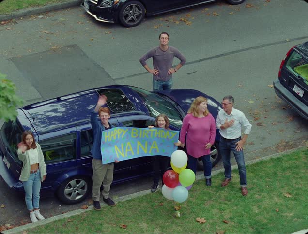 A family stands in a yard beside their vehicle and holds up a sign that says happy birthday nana