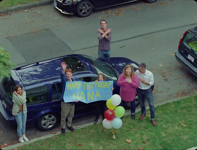 Family stands outside house with happy birthday nana sign waving and smiling