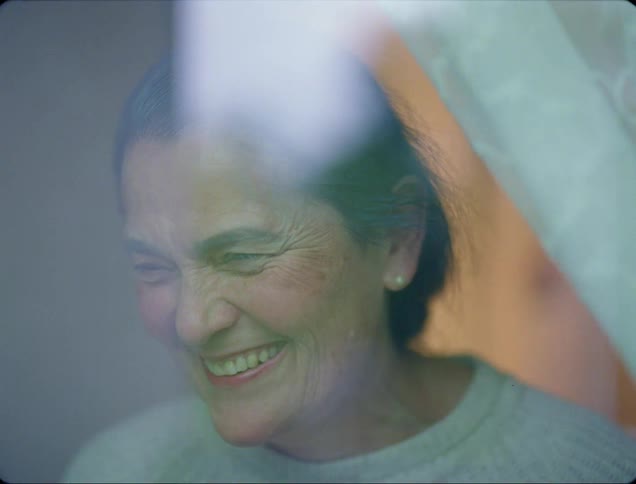 A graying woman blows kisses as she laughs and talks while standing near a window