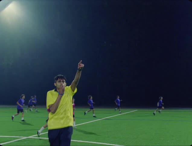 A referee on a soccer field blows his whistle and puts his arm up and runs as players continue running on the field