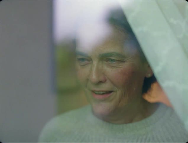 An elderly woman moves a curtain to look out of her window before laughing and waving at what she sees outside