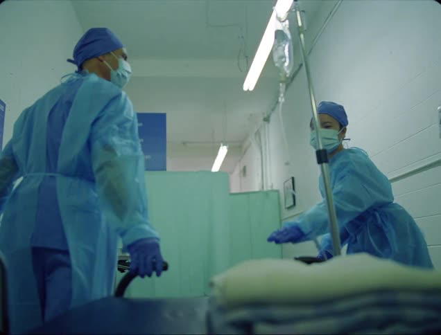 A man with gloves a mask and scrubs passing a stretcher to a woman in a hospital