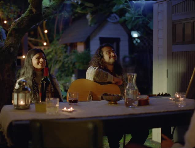 A man holding a guitar while sitting at a table with a pair of women nods and smiles at someone out of frame before the smile fades from his face