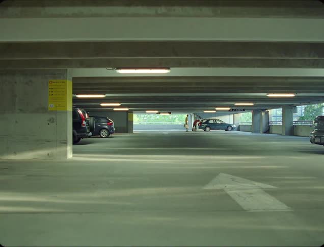 A pair of people carry bags through a parking garage to a parked car and place the bags in the rear of the vehicle