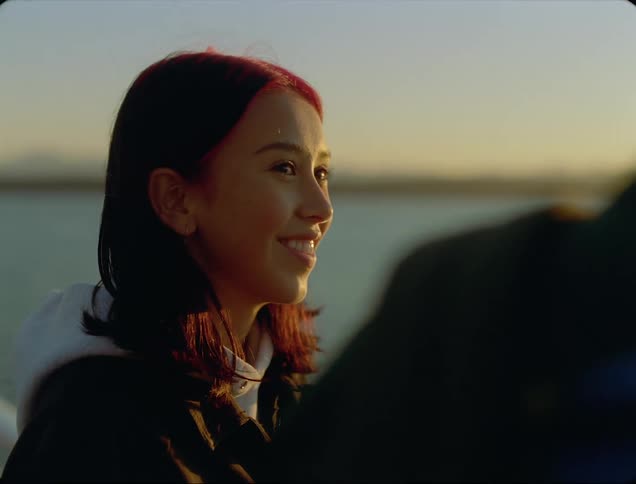 A young asian woman smiles as she talks with another person while standing at a waterfront in the evening