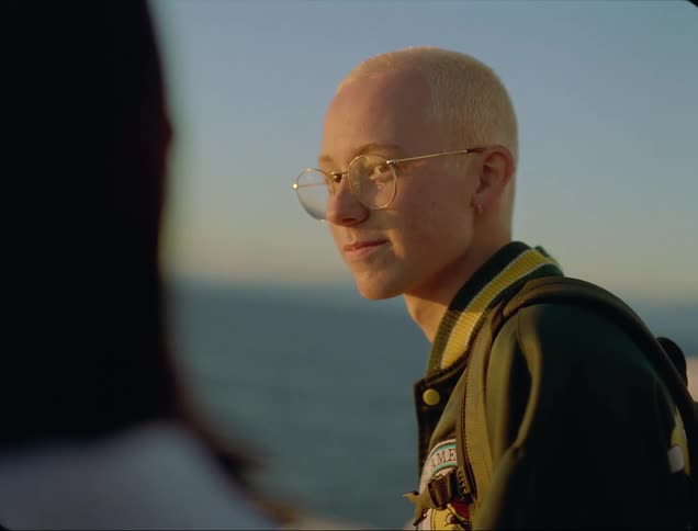 A man with glasses a jacket and a backpack standing with the ocean and sky in the background
