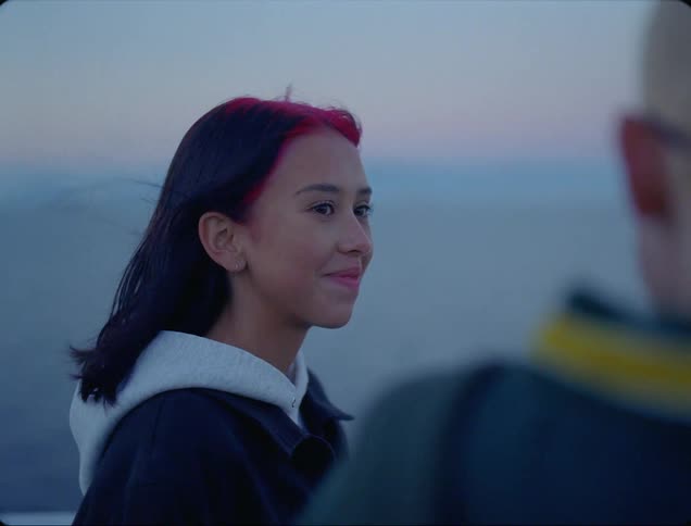 A teenage girl with pink and black hair smiles at her male friend as they stand outside overlooking the ocean