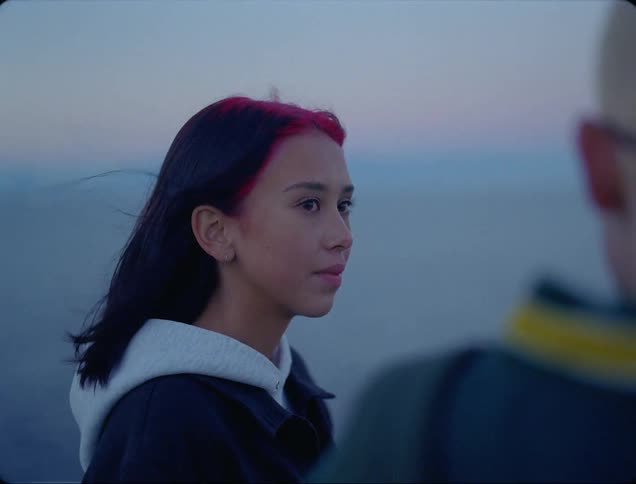 A teenage girl standing outside looks at the boy next to her and smiles