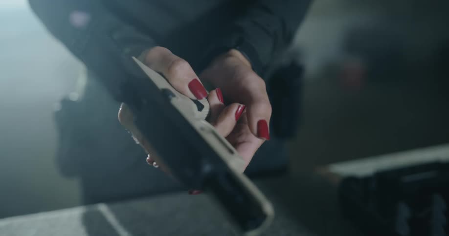 A woman takes a pistol from a table covered in firearms and loads it before lifting it to take aim