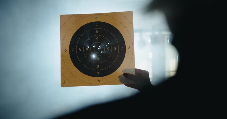 A marksman in a misty room holds a paper shooting target up to the light to see the holes