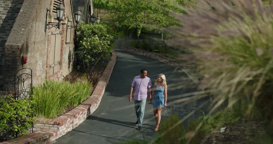 A woman and man hold hands while walking down a narrow road between a building a hill