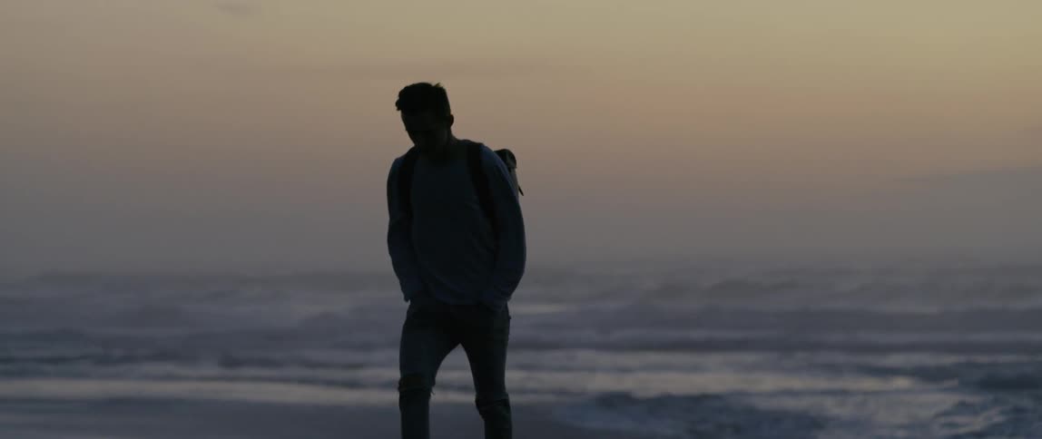 A man wearing a backpack walks away from the shore on a beach at sunset