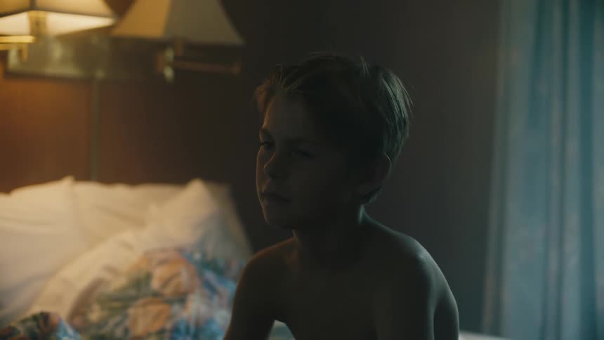 Shirtless child sits on a bed in his room