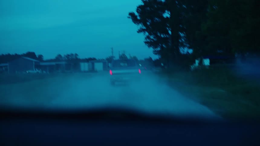 Vehicle follows another vehicle past buildings along dirt road throwing up dust as it approaches cross road