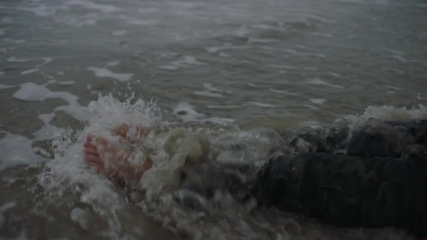 A wave crashes over a clothed person sitting on a beach