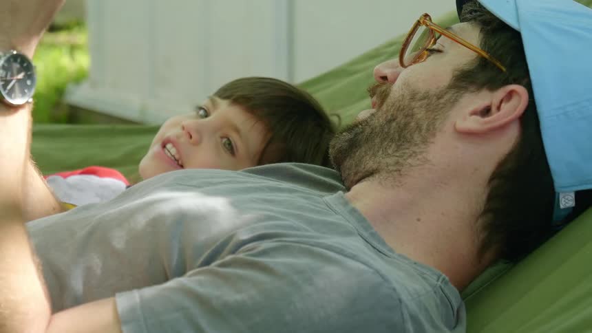 Father with a blue cap glasses and a beard lies in a hammock with his son in the garden