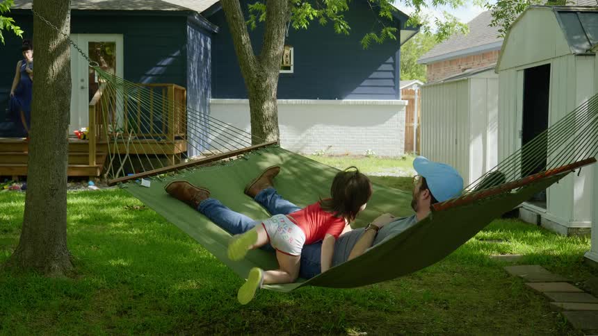 A man with a hat and glasses laying on a hammock with a boy crossing over him with a woman holding a baby in the background