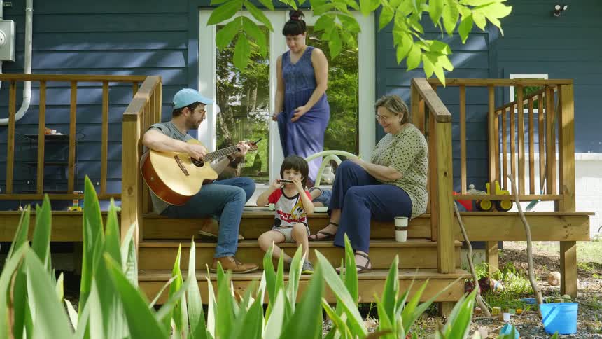 A boy plays the guitar while his father plays the guitar as they sit on their porch with the family