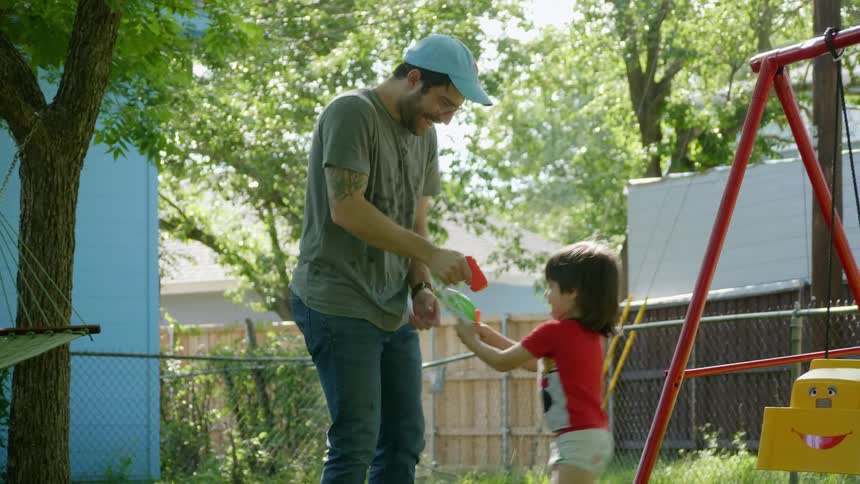A man and a boy playfully spray each other with water guns in the back yard of a house during the day