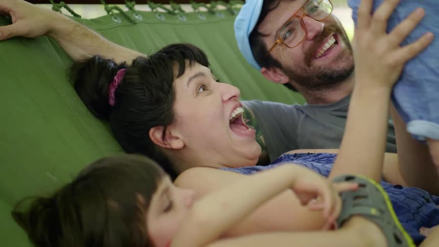 A woman lying on a hammock with her husband lifts their baby up in the air and talks to their toddler
