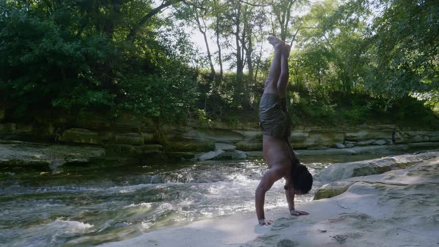 A young man does a hand stand next to a river