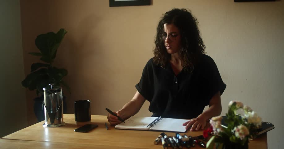 A woman sketches with markers in a notebook while sitting at her kitchen table with coffee