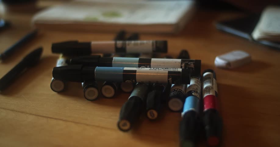 A woman switches markers from a pile of them on a desk while drawing in a sketchbook