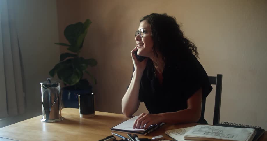 Woman sitting at wooden table with sketchbook and pens nearby talks on mobile phone