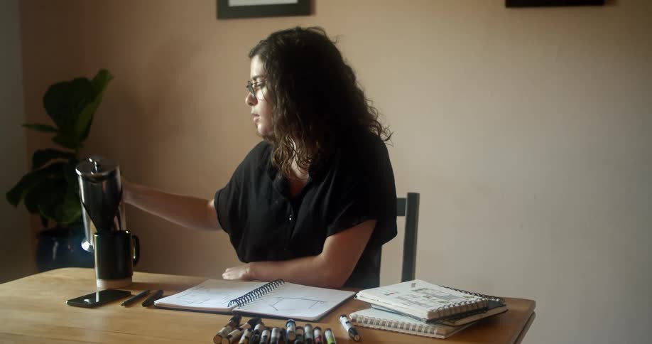 A woman pours herself some coffee from a french press as she sits at a table with some notebooks