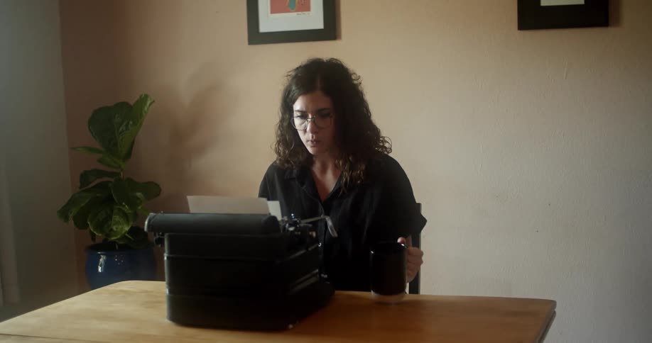 A woman sitting at home types on a vintage typewriter while drinking from a mug
