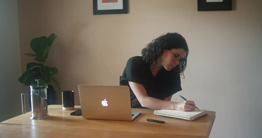 Woman sitting at wooden table views laptop and makes notes