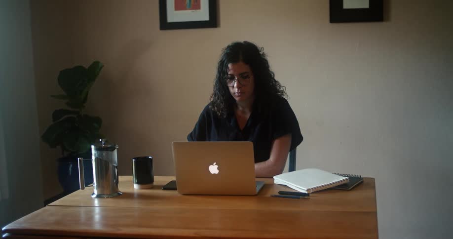 A woman sitting at a table drinks from a coffee mug before turning to look at the laptop on the table before her