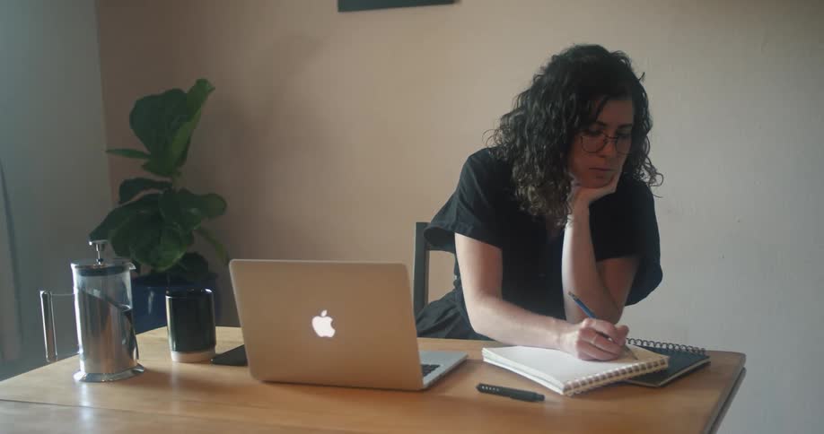 A woman sitting at a desk turns away from her laptop to write in a notebook