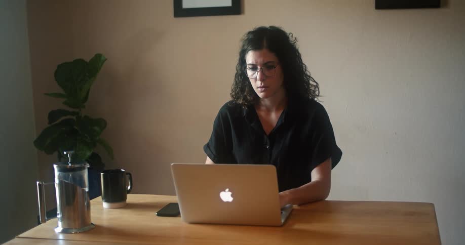 Woman wearing eyeglasses sits at wooden table working on laptop