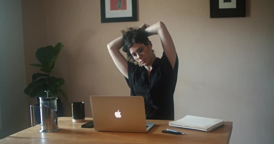 A woman sitting at a desk while typing on a laptop stops to stretch as she reads the laptop screen