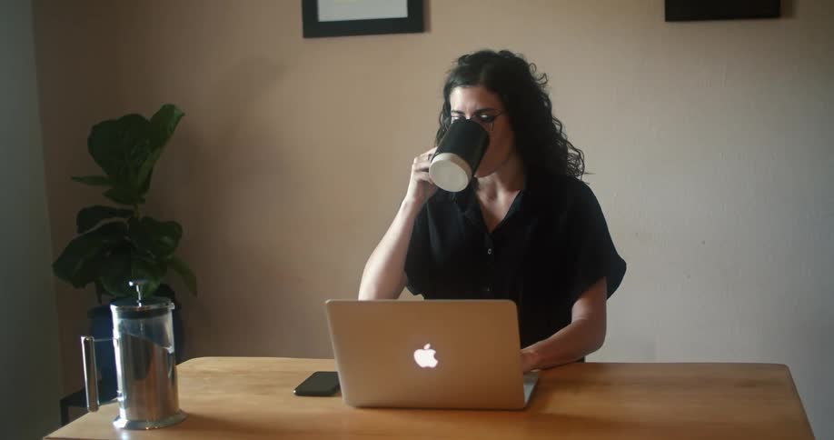 A woman takes a sip from a coffee mug while typing on a laptop at a table