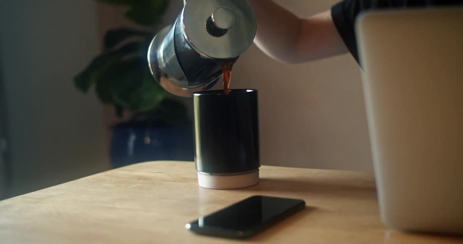 A person sitting in front of a laptop at a table pours coffee from a tall coffee pot into a mug