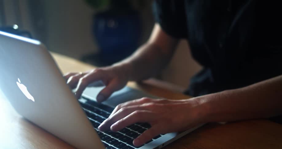 Woman with curly hair and glasses sitting at a table drinks a cup of coffee and works on her laptop in a room
