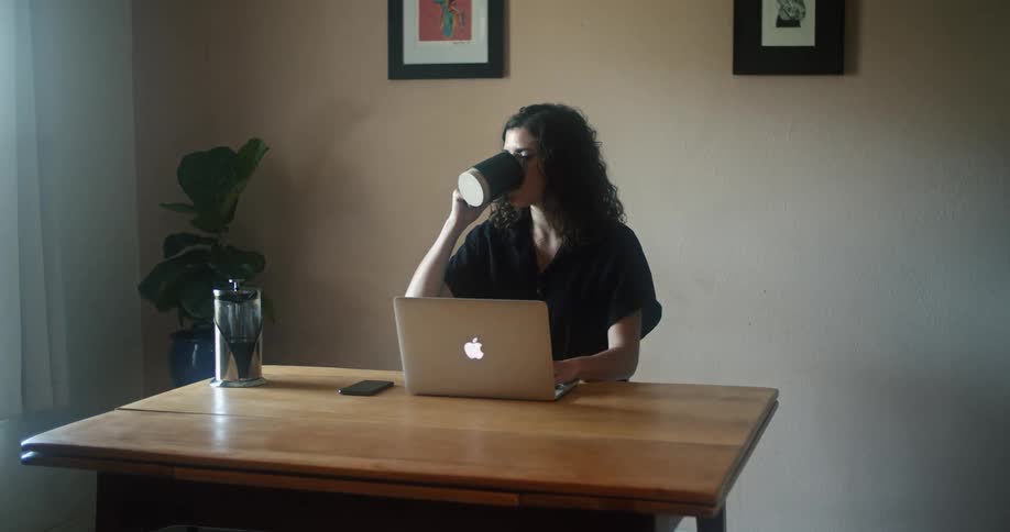 A woman sits at a small table with a laptop and takes a drink from a mug