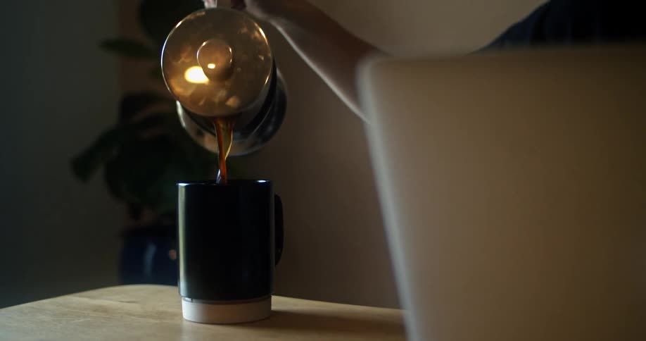 A woman at home sits with her laptop open and pours herself a cup of coffee