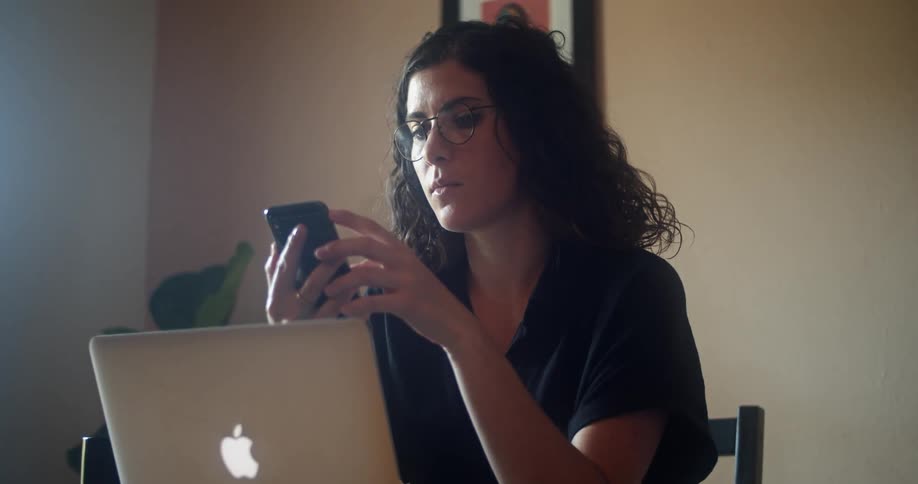 A woman sits at a table in front of a laptop and looks at her cell phone