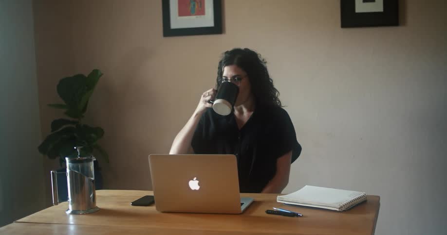 A woman sitting at table takes a sip from a coffee mug while looking at the screen of a laptop