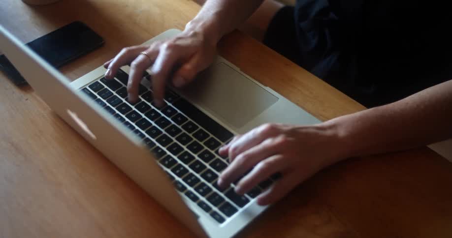 A person sitting at a table with a smart phone typing on a laptop