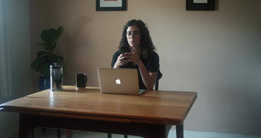 Woman with glasses and a black blouse sitting at a table works on her laptop and checks her smartphone in a room