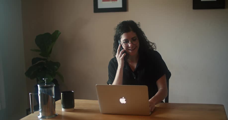 A woman sits at a desk with a laptop and answers her cell phone